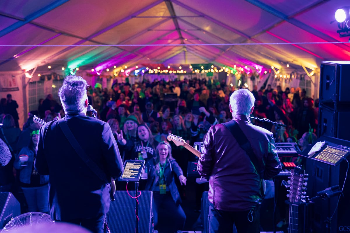 back of band members playing on a stage in front of a large crowd under a tent