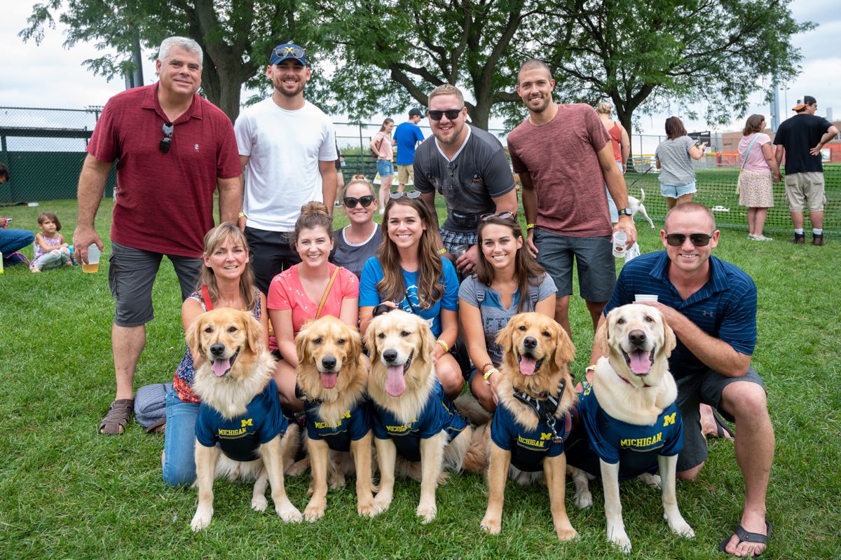 four golden retrievers and a yellow lab wearing Michigan football jerseys posing with their owners at Royal Oak's Barktoberfest