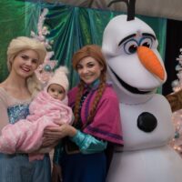 Anna, Elsa, and Olaf posing with baby wrapped in pink blanket and pink hat during Royal Oak Jingle