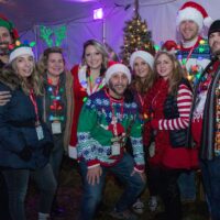 group of friends in Christmas outfits and Christmas light necklaces and headbands under large white tent during Wyandotte's Santa Pub Crawl