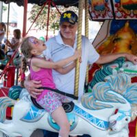 man in University of Michigan hat holding little girl with butterfly face paint on carousel at Wyandotte's Swiggin' Pig