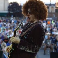 closeup of guitar playing wearing afro wig with crowd in the background at Wyandotte's Street Art Fair