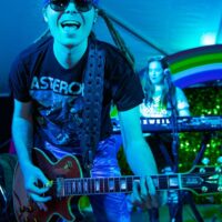 band guitar playing wearing sunglasses on stage singing to camera with rainbow backdrop