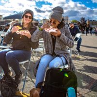 two women sitting on folding chairs wearing sunglasses and eating tacos