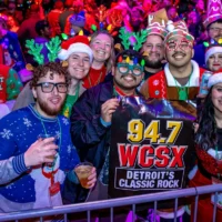 Crowd of people wearing Christmas sweaters, funny hats, and glasses and holding a 94.7 WCSX Detroit's Classic Rock banner.