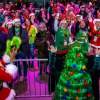 Crowd of people wearing Christmas sweaters and fun themed outfits cheering at someone dressed as a Christmas tree.