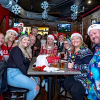 Crowd of people wearing Christmas sweaters and fun themed outfits at a bar.