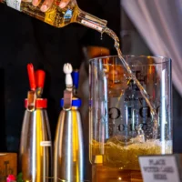 A clear bottle of amber liquid is being poured into a large glass dispenser, causing the liquid to foam and splash. Two metal dispensers are visible on the bar counter.