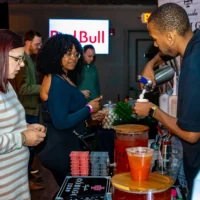 A man working at an event bar uses a metal canister to dispense a foam topping onto a small cocktail being served to two waiting female guests.