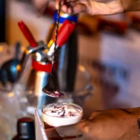 A close-up of a mixologist using a twisted bar spoon to sprinkle dried red petals onto a cocktail topped with creamy white foam.