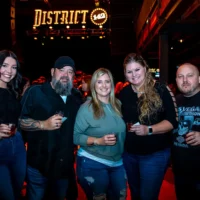 Five smiling event attendees holding drinks pose together in front of a brightly lit "DISTRICT 142" sign in a dark venue.
