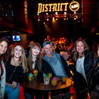 A group of friends smiling around a small table with cocktail samples, enjoying a show at the District 142 music venue. A band performs on the stage in the dark background.