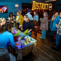 A lively group socializes at the Detroit River Cocktail Showdown. Attendees gather around a table where a woman prepares citrus garnishes, with the "District 142" sign glowing in the background.