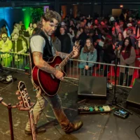 A male musician with tattoos and dark hair wears a vest while playing a dark red acoustic guitar on stage for a large crowd separated by a metal barrier.