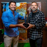 A man in a blue shirt and a man in a plaid shirt smile happily while holding a silver cocktail shaker trophy together at an event.