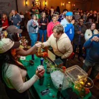 A server hands a sample drink to an attendee over a green bar setup, while another server prepares a cocktail in the foreground of a crowded social event.