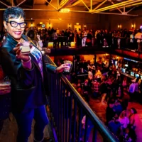 A woman smiles brightly, holding up two cocktails from a balcony at the crowded Detroit River Cocktail Showdown. Guests fill the lower level of the dimly lit, energetic venue.