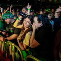 Two women in green at a crowded St. Patrick's Day event laugh and whisper together. The woman on the left smiles widely and holds up a cell phone as the surrounding crowd cheers with their hands raised.