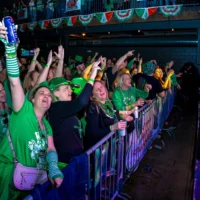 A jubilant crowd of attendees, all wearing green for St. Patrick's Day, cheers and raises their hands and drinks at a lively indoor concert.