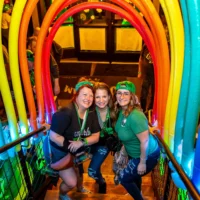 Three women smiling brightly pose on a staircase, wearing green clothing and festive beads under a colorful rainbow arch.