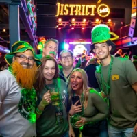 A festive group of six people wearing green beads and hats, including a man with a fake beard, smiles while holding drinks at a crowded St. Patrick's Day party.