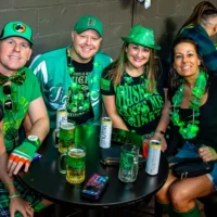 Four people wearing green St. Patrick's Day attire smile happily while gathered around a table with drinks in a bar.