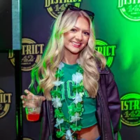 A smiling woman with blonde hair at a party, wearing a green top, dark jacket, and green shamrock leis, holding a drink.