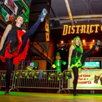 Two energetic female dancers in black and brightly colored traditional costumes perform synchronized high kicks on a green-lit stage.