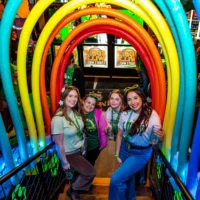 Four happy women in green St. Patrick's Day outfits stand on stairs, framed by a brightly lit rainbow arch in a venue.