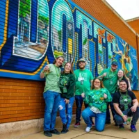 A cheerful group of seven people wearing green, holding beer mugs, pose for a photo in front of a large, colorful city mural.