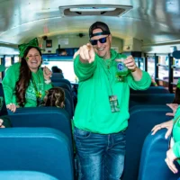 A man in a green hoodie, sunglasses, and a cap smiles broadly and points at the camera, holding a green cocktail on a party bus. A laughing woman with a large green bow stands behind him.