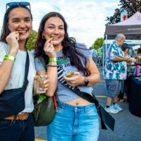 Two women with wide smiles sample pickles at a crowded outdoor festival. They wear wristbands and casual clothing, with event tents visible behind them.