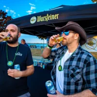 Two attendees at an outdoor beer festival sample drinks under a Warsteiner tent at dusk. One man wears a cowboy hat and sunglasses.