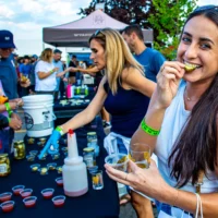 A woman smiles while biting a pickle sample at an outdoor festival, holding a glass of beer as a vendor serves small cups of sauces and pickles from a black table.