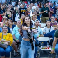 A woman wearing a 'Birthday Girl' sash throws a pickle toward the camera while standing in front of a large, seated crowd.