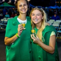 Two women, both smiling, wear matching bright green novelty costumes and hold Wyandotte Beer Fest sample glasses at an outdoor event.