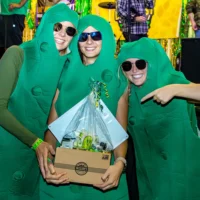 Three people in bright green pickle costumes pose with sunglasses, smiling as they show off a gift box of McClure's Pickles.