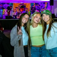 Three women smiling happily pose together in front of a brightly lit concert stage with a band playing. The woman on the left makes a peace sign.