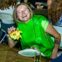 A blonde woman in a vibrant green pickle costume, wearing clear glasses, laughs loudly while taking a bite of a slice of pizza.