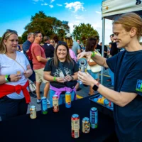 A vendor pours a sample drink from a can into a glass for two female attendees at an outdoor event. One woman smiles brightly while watching the pour.