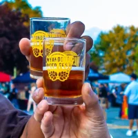 Two hands hold up two small tasting glasses filled with amber beer. Each glass features the yellow "Wyandotte Beer Fest" logo, set against a blurry outdoor festival background.